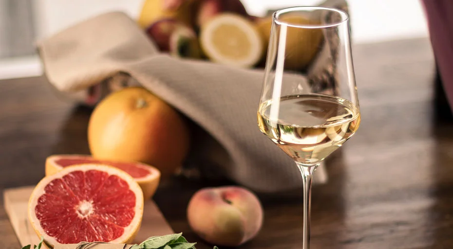 Weinglas der Serie Sky Sensis plus von Eisch mit Weißwein neben Grapefruit, Pfirsichen und Zitronen auf Holztisch. 