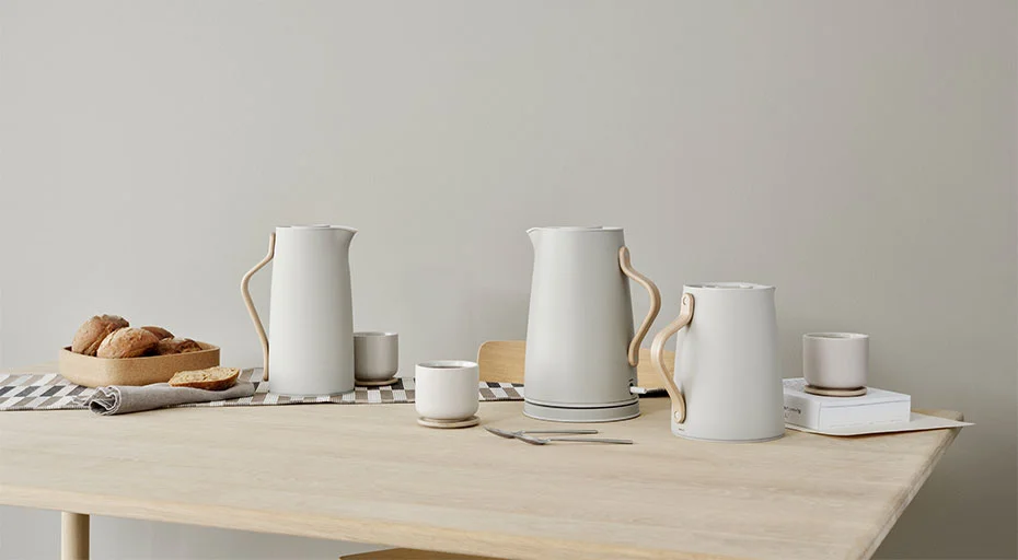 Minimalistische Tischszene mit Stelton Wasserkochern, Tassen, einem Tablett mit Brot und einem karierten Tischläufer auf hellem Holztisch.