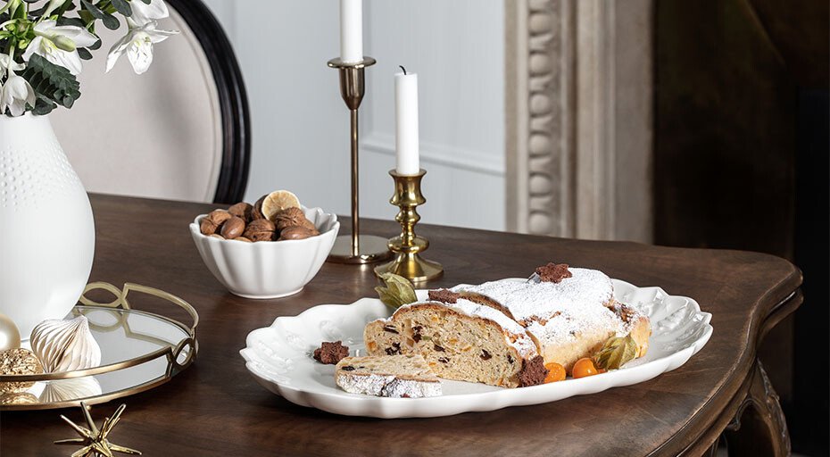 Tisch mit Stollen auf  weihnachtlicher Servierplatte der Serie Toy's Delight Royal Classic von Villeroy &amp; Boch, neben Dekorkerzen, Vase mit Blumen und Schale mit Nüssen. Elegante Weihnachts-Tischdekoration in klassischem Stil.