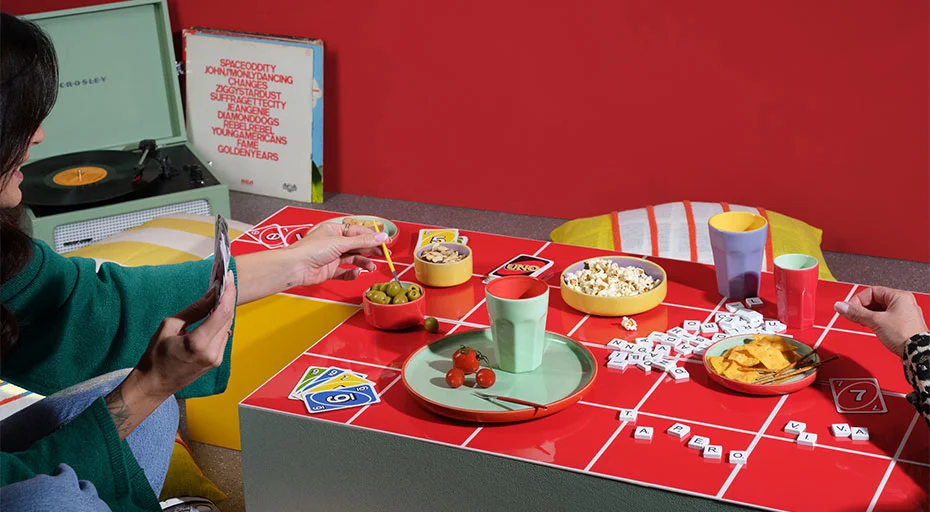 Geschirr von ASA Tapero &amp; Ti amo viva: Personen spielen ein Brettspiel auf einem rot-karierten Tisch, umgeben von Snacks und Getränken. Plattenspieler im Hintergrund.