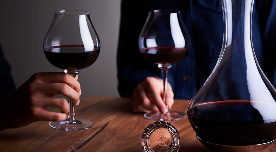 Zwei Personen halten Weingläser, eine Dekantierkaraffe steht auf dem Tisch. Im Hintergrund dunkler, unscharfer Hintergrund.