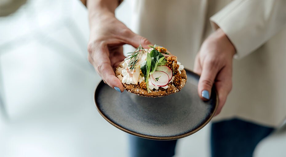 Person hält einen Brot-Teller von Villeroy &amp; Boch mit einem belegten Brot, garniert mit Radieschen, Kräutern und Sprossen.