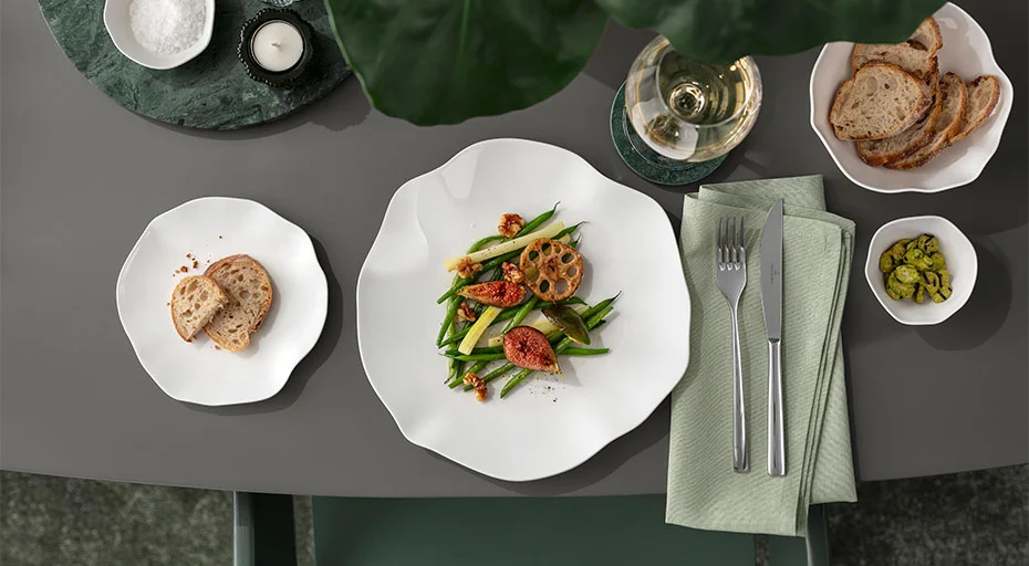 Speiseteller Villeroy &amp; Boch Pura mit edlem Gericht neben Brotteller der gleichen Serie auf hochwertig gedecktem Tisch neben Besteck und einem Glas Weißwein.
