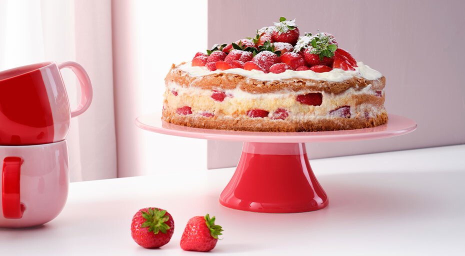 Erdbeertorte auf roter Kuchenplatte der Serie Joy von ASA, dekoriert mit Sahne und Beeren, neben zwei gestapelten roten Tassen der selben Serie und Erdbeeren.