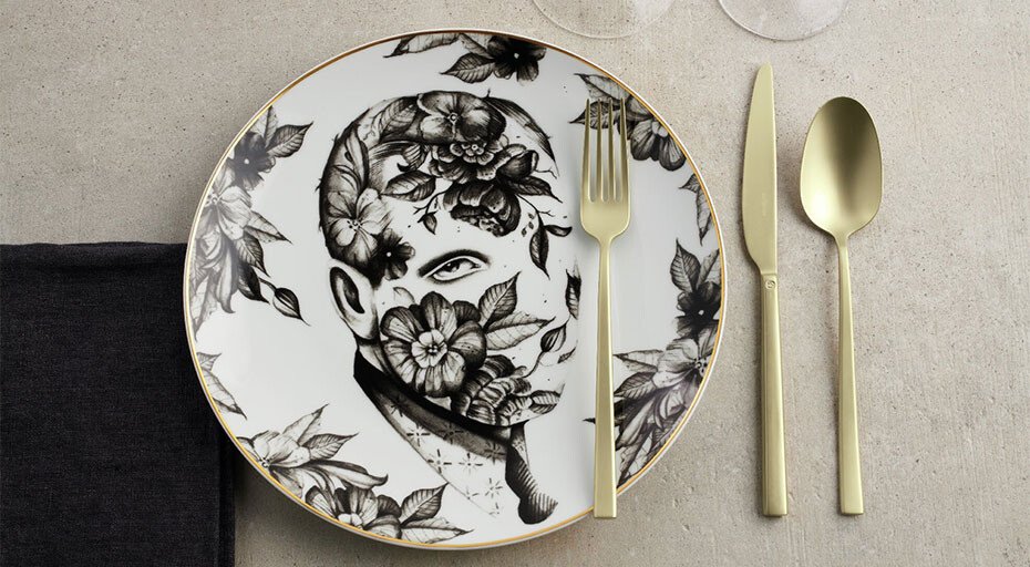 Teller mit floralem Muster, goldfarbenes Besteck, schwarzes Geschirrtuch, auf grauem Untergrund.