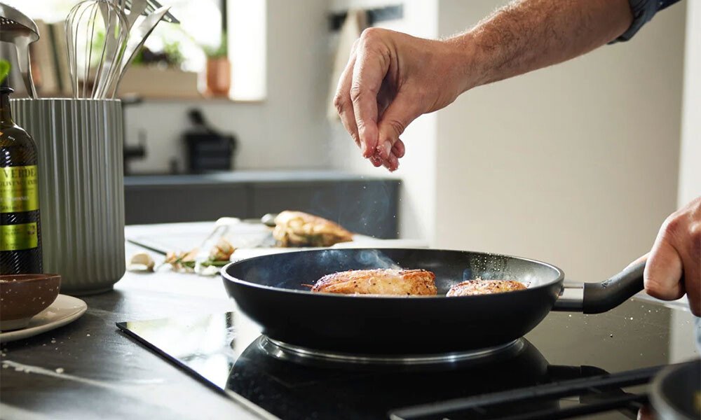 Person würzt in einer Küche Fleisch in einer Essential-Pfanne von Fissler auf einem Herd. Küchenutensilien und Ölflasche im Hintergrund.