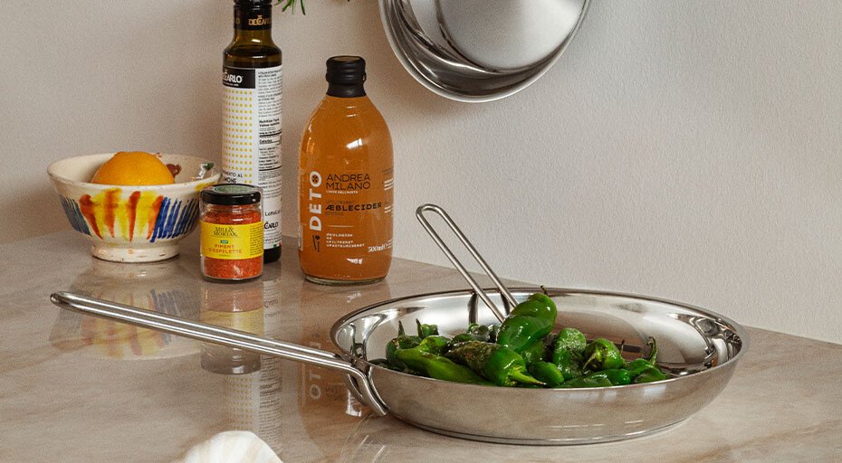 Eine Pfanne von Eva Solo mit grünen Paprikaschoten auf einem Küchentresen, daneben Gewürze und eine Flasche Saft.