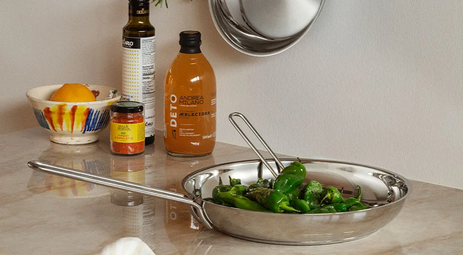 Eine Pfanne von Eva Solo mit grünen Paprikaschoten auf einem Küchentresen, daneben Gewürze und eine Flasche Saft.