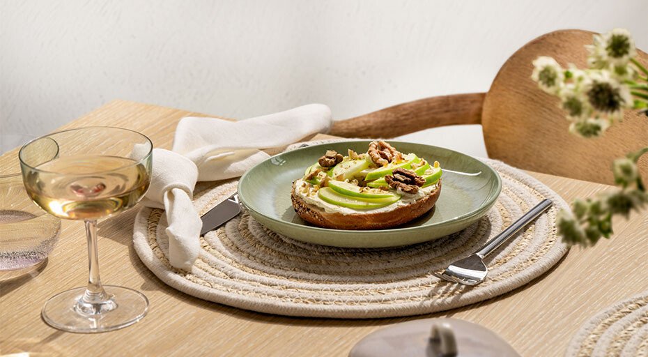 Bagel mit Avocado, Nüssen und Apfelscheiben auf grünem Teller, daneben ein Weinglas der Serie Like Pearl von Villeroy &amp; Boch auf einem gedeckten Holztisch.