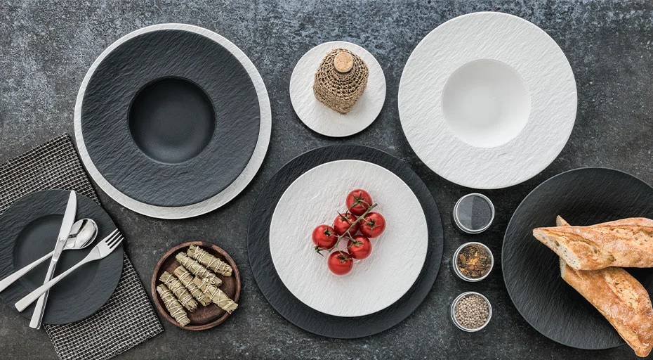 Elegante Tischdekoration mit schwarzen und weißen Tellern der Serie Manufacture Rock von Villeroy &amp; Boch, Tomaten, Brot und Gewürzen auf grauem Untergrund.