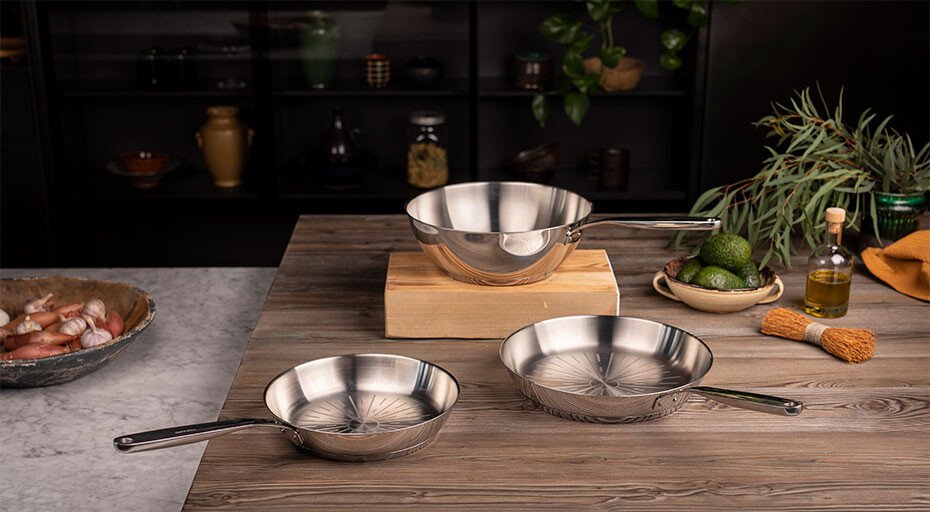 Drei Edelstahlpfannen von Fiskars auf einem Holztisch in einer Küche, umgeben von Kräutern und Gewürzen.