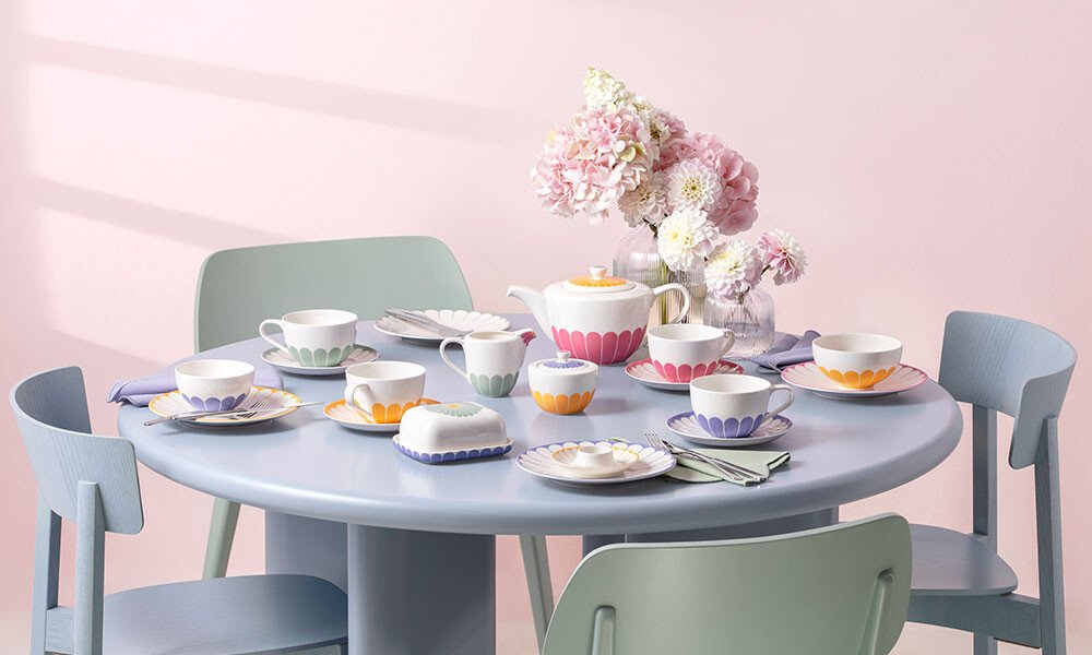 Runder Tisch mit bunter Teeservice-Garnitur der Serie Fleur von Villeroy &amp; Boch, Pastellstühlen und einem Blumenarrangement in einer Vase.