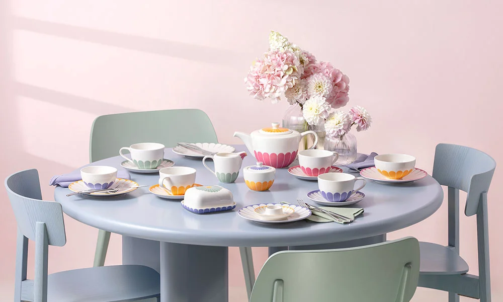 Runder Tisch mit bunter Teeservice-Garnitur der Serie Fleur von Villeroy &amp; Boch, Pastellstühlen und einem Blumenarrangement in einer Vase.