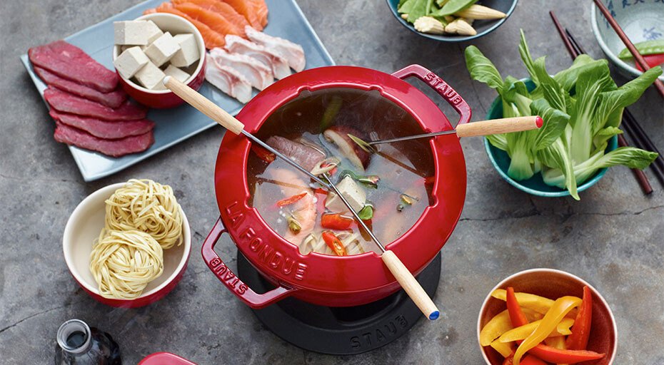Rotes Fondue-Set mit Brühe, umgeben von rohem Fleisch, Gemüse, Tofu und Nudeln auf einem Tisch.