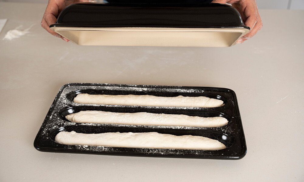 Baguetteteig-Rohlinge in einer speziellen Backform, bereit zum Gären. Hände halten einen Blechdeckel darüber.