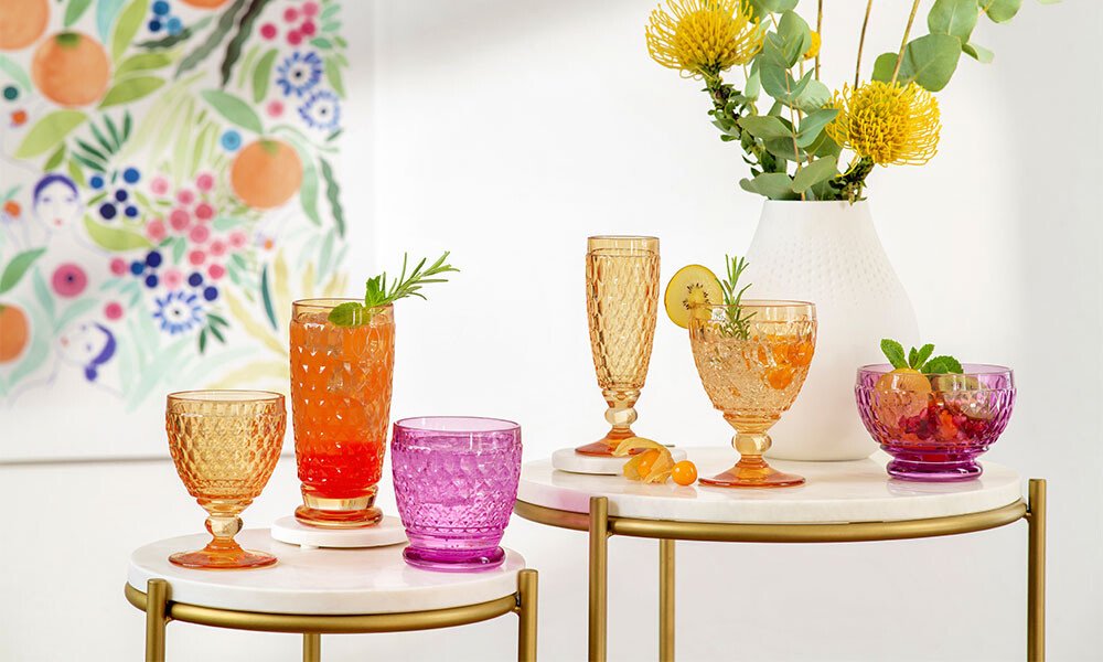 Mehrfarbige Gläser der Serie Boston von Villeroy &amp; Boch mit Getränken und Zitronenscheiben auf runden Tischen, daneben eine Vase mit gelben Blumen.
