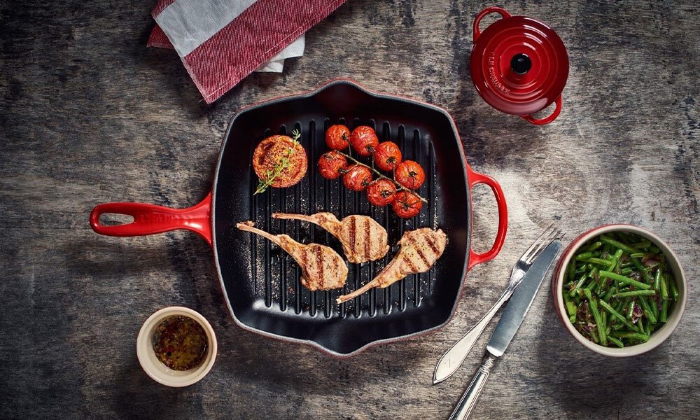 Grillpfanne von Le Creuset mit Lammkoteletts, gerösteten Tomaten und Kräutern, umgeben von Beilagen in Schalen und Besteck auf grauem Tisch.