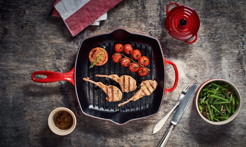 Grillpfanne von Le Creuset mit Lammkoteletts, gerösteten Tomaten und Kräutern, umgeben von Beilagen in Schalen und Besteck auf grauem Tisch.