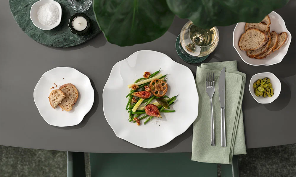 Tisch mit elegant angerichtetem Teller der Serie Pura von Villeroy &amp; Boch, Brot, Besteck, Glas Weißwein und kleinen Schalen mit Beilagen auf grauer Tischplatte.