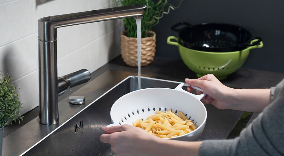 Person spült gekochte Nudeln in einem Sieb von Trebonn unter einem Wasserhahn ab, Grünes Sieb im Hintergrund.