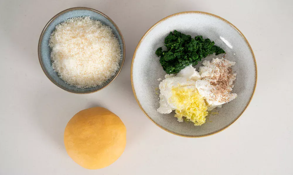 Schüsseln mit geriebenem Käse, Spinatmischung, Ricotta, Zitronenschale und Teigkugel auf weißem Hintergrund.
