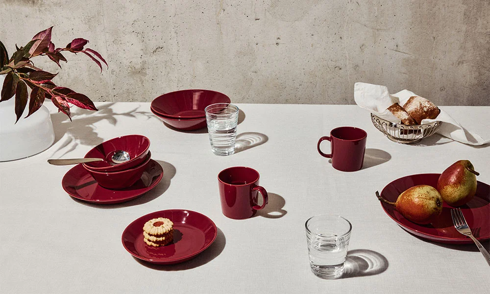 Minimalistisch gedeckter Tisch mit rotem Geschirr der Serie Teema von Iittala, Wasser, Birnen und Gebäck auf weißem Tischtuch vor heller Wand.