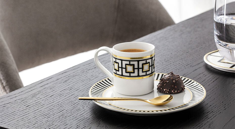 Espressotasse der Serie MetroChic von Villeroy &amp; Boch mit gold-schwarzem Muster, Schokoladentrüffel und goldener Löffel auf dem Teller, Tisch aus dunklem Holz.