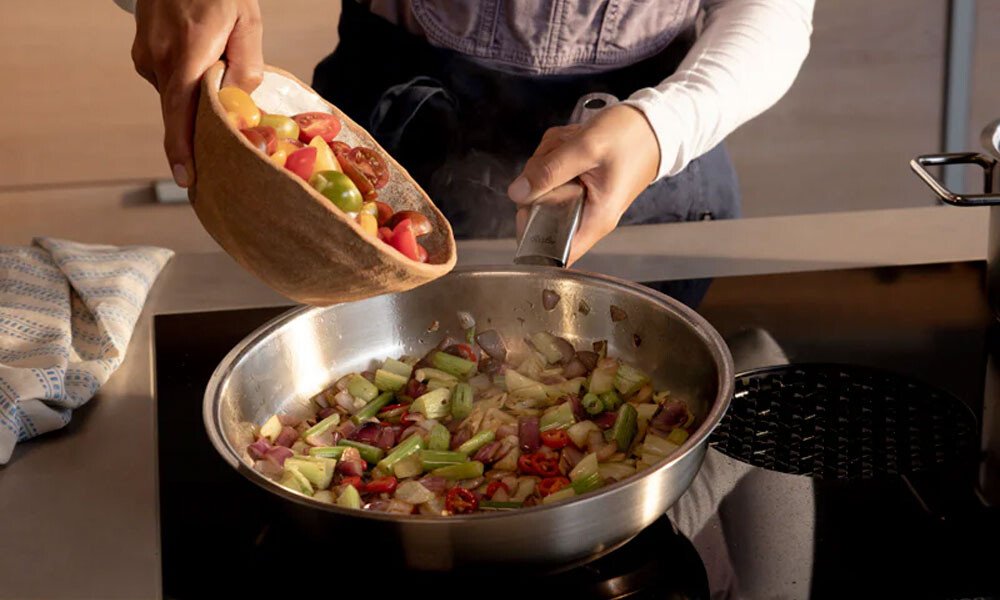 Person gibt geschnittenes Gemüse aus Holzschüssel in Pfanne der Pure Collection von Fissler auf Herd, um eine bunte Pfannengericht zuzubereiten.