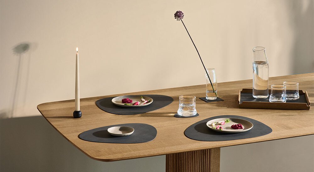 Eleganter Tisch mit Tischsets und Untersetzern, Geschirr, Gläsern und minimalistischer Blumendekoration. Skandinavisches Design mit Holzoberfläche.