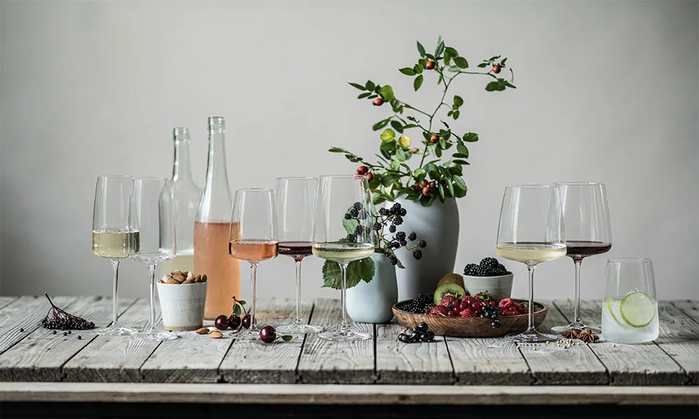 Stillleben mit Weingläsern, Flaschen, Schalen mit Beeren, Nüssen und Vase auf Holzoberfläche.