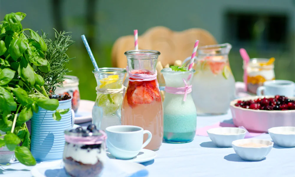 Ein Tisch mit Getränken in Glaskaraffen, frischen Kräutern, Dessertgläsern und Beeren in Schalen, im Freien gedeckt.
