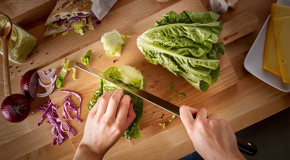Hände schneiden mit einem Messer der Serie Gourmet von Wüsthof Römersalat auf einem Holzbrett, umgeben von geschnittenem Rotkohl, Zwiebeln und Käse.