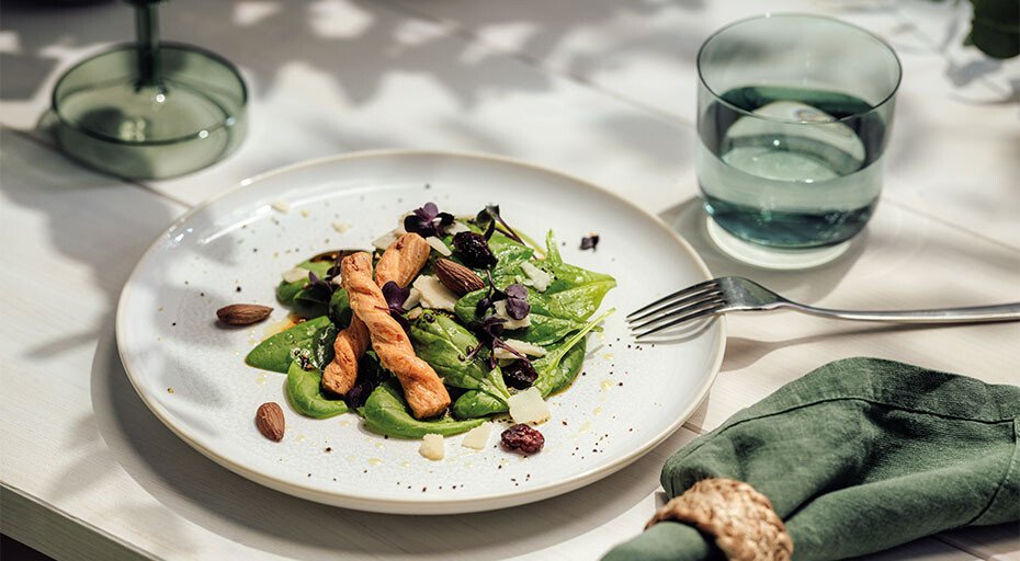 Teller der Serie Crafted Cotton von Villeroy &amp; Boch mit grünem Blattsalat, Mandeln, Rosinen und Croutons. Daneben Glas Wasser, Gabel und grüne Serviette auf hellem Tisch.