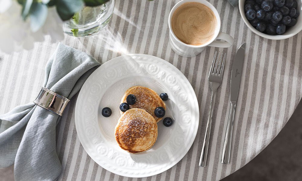 Weißer Teller er Serie Cellini von Villeroy &amp; Boch mit zwei Pfannkuchen und Blaubeeren, daneben Kaffee und Besteck auf gestreiftem Tischtuch.