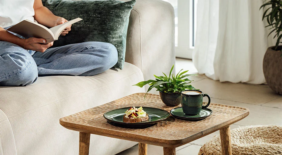 Person liest auf Sofa, daneben ein Holztisch mit Tasse, Teller der Serie Lave vert von Villeroy &amp; Boch und Pflanze in gemütlichem Wohnzimmerambiente.