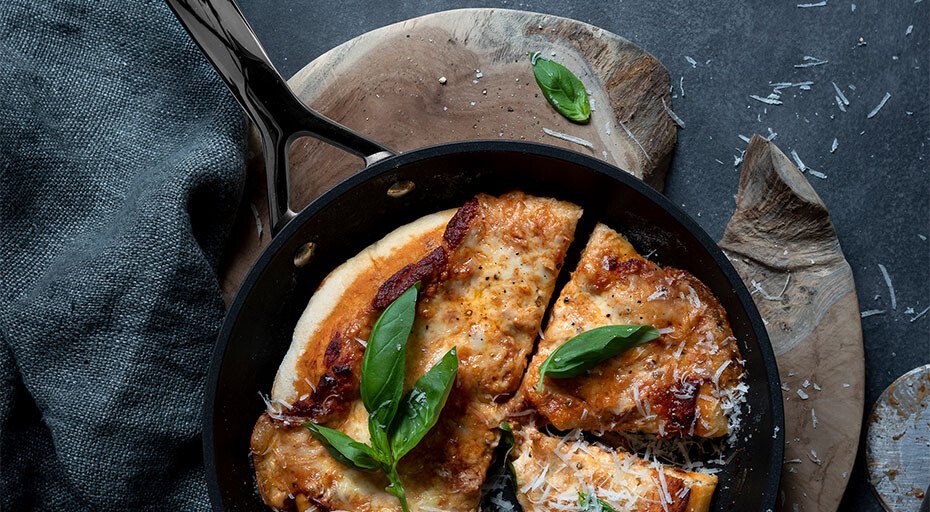 Pizza in einer Pfanne der 3Ply-Serie von Le Creuset mit Basilikum und geriebenem Käse, auf Holzbrett serviert, dunkler Hintergrund.