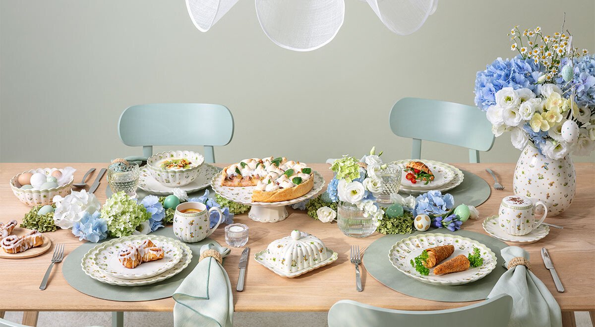 Festlich gedeckter Oster-Tisch mit Frühlingsdekoration, Blumenarrangements, Porzellantellern der Serie Easter Delight von Villeroy &amp; Boch und diversen Speisen unter einer weißen Lampe.