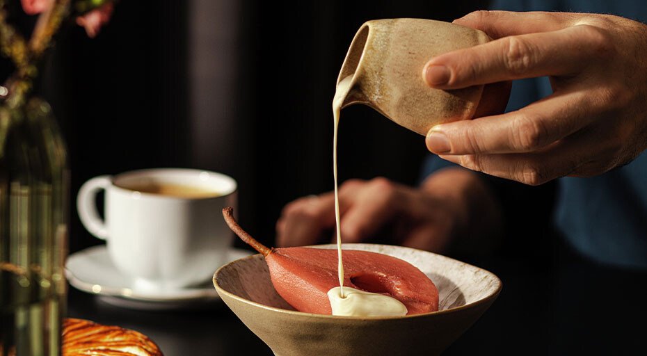 Person gießt Soße über pochierte Birne in Schale, im Hintergrund eine Tasse Kaffee und ein Gebäckstück.