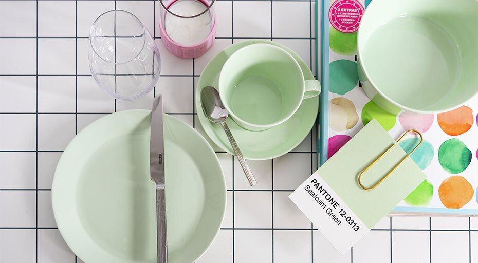 Pastellgrünes Geschirr der Serie Tric Lime Cream von Thomas auf kariertem Tisch, mit Besteck, Glas, Pantone-Karten und Kerze in Rosa.