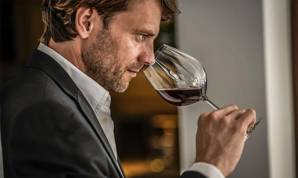 Ein Mann im Anzug prüft konzentriert ein Glas Rotwein in einem hellen Raum.