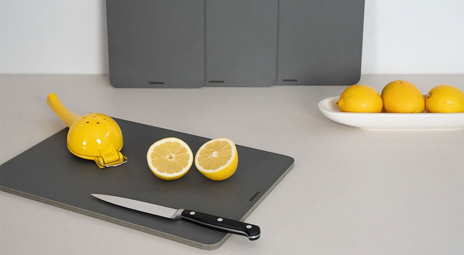 Zitronenhälften, Messer und Zitronenpresse auf spülmaschinenfestem Schneidebrett; Schale mit Zitronen im Hintergrund.