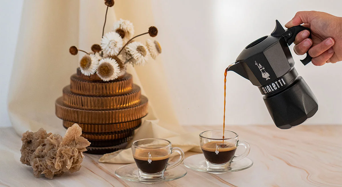 Gießen von Espresso aus einem Espressokocher von Bialetti in zwei Gläser, dekoriert mit getrockneten Blumen auf einem Tisch.