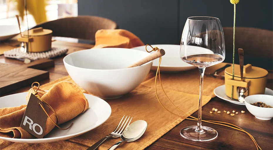 Teller und Schale der Serie TAC Gropius weiß von Rosenthal auf Elegant gedecktem Tisch mit Weinglas, Goldbesteck und orangefarbenen Akzenten auf Holztischoberfläche.