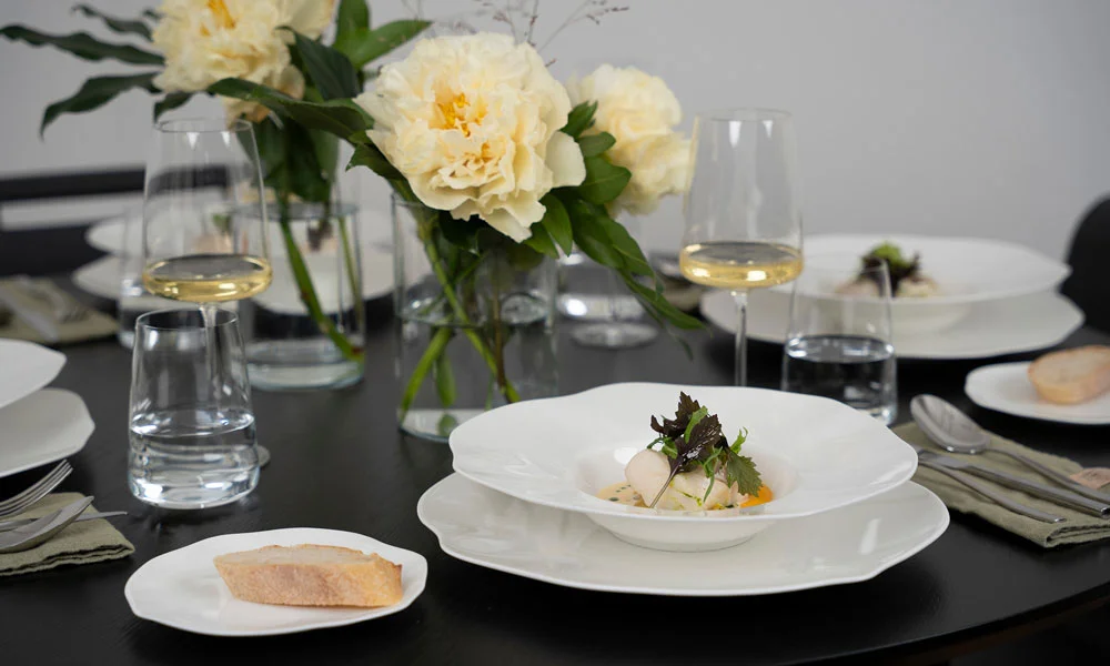 Gourmetteller der Serie Pura von Villeroy &amp; Boch mit fein angerichtetem Gericht, Blumen und Weinglas auf schwarzem Tisch, stilvolles Tischgedeck.