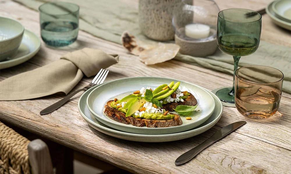 Avocado-Toast auf Villeroy &amp; Boch Perlemor Teller, gedeckter Tisch mit Besteck, grüne Gläser, rustikaler Holztisch, natürliche Dekoelemente.