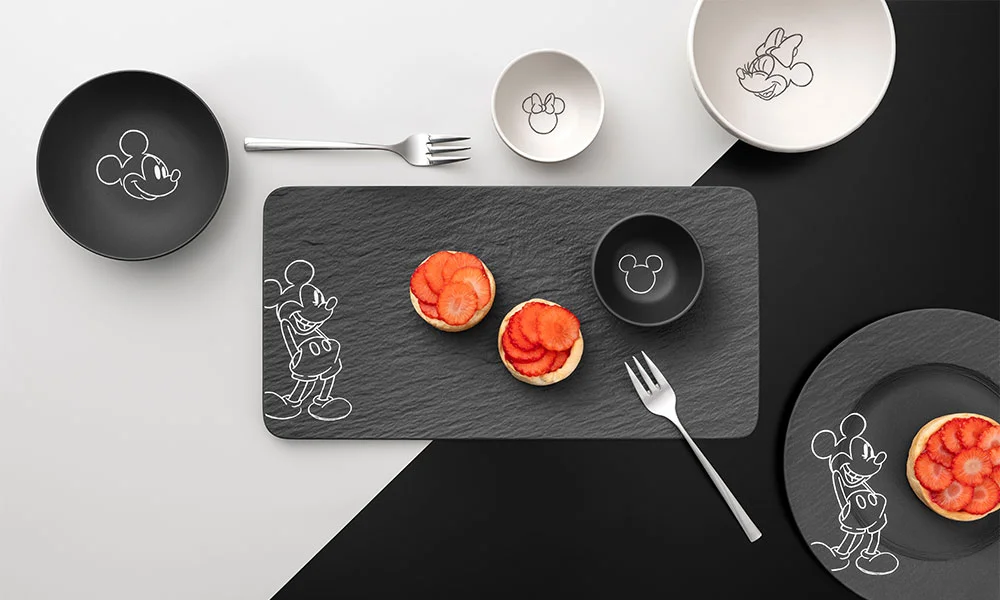 Teller- und Schüsselset der Serie Villeroy &amp; Boch Manufacture Rock – Mickey Mouse mit Mickey-Maus-Motiven, schwarze und weiße Designs, serviert mit Gebäck und Erdbeeren auf Schieferplatte.
