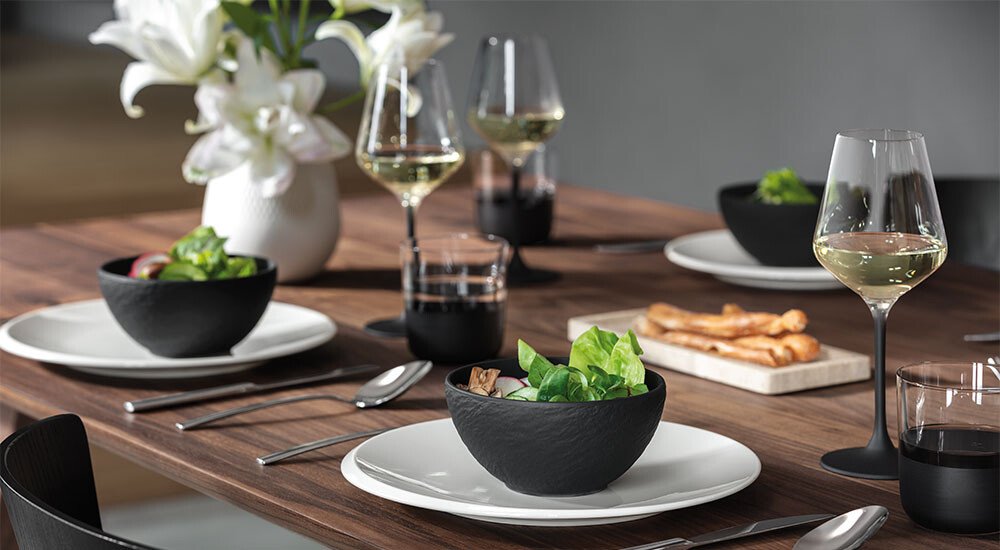 Moderner Esstisch mit schwarzen Schalen, Gläsern und weißem Geschirr, dekoriert mit Blumen und Brotstangen.