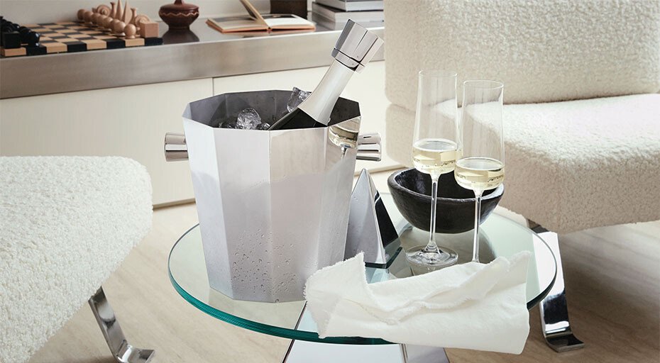 Silberner Weinkühler mit Flasche auf Glastisch, zwei gefüllte Sektgläser der Serie Eugenia von Alessi daneben, im modernen Wohnzimmerambiente.
