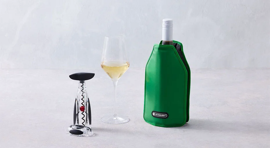 Flasche im grünen Le Creuset Kühlmanschettenzug, Glas Weißwein und Korkenzieher auf hellem Untergrund.