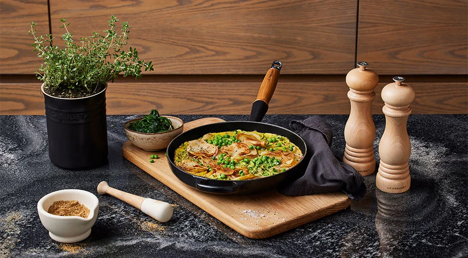 Gusseisenpfanne von Le Creuset mit Pfannengericht mit Fleisch und Gemüse, dekoriert mit Kräutern, auf Holzbrett. Kräutertopf, Gewürzschälchen und Pfeffermühlen daneben.